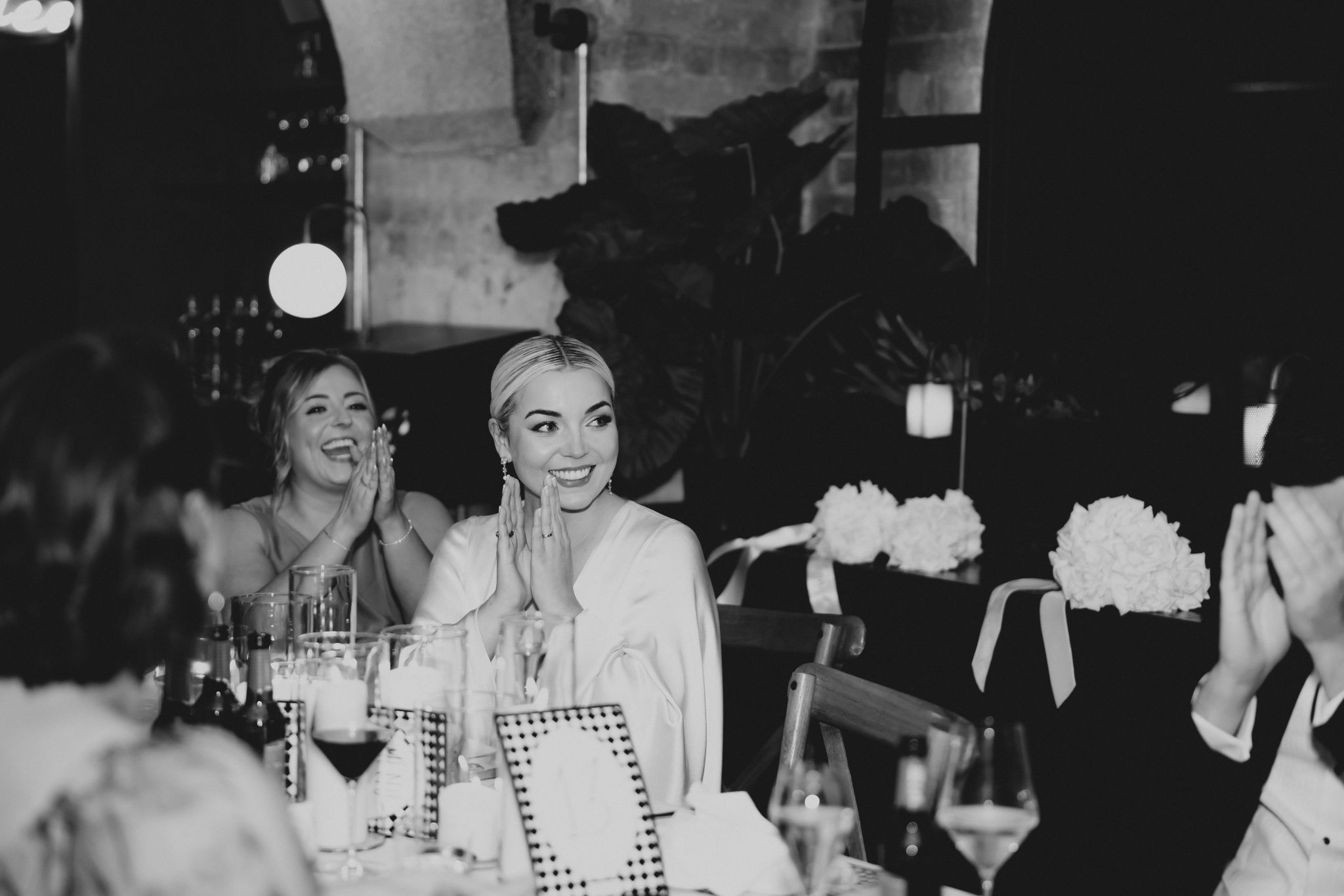 Bride seated at wedding table wearing white dress with flowers in background.