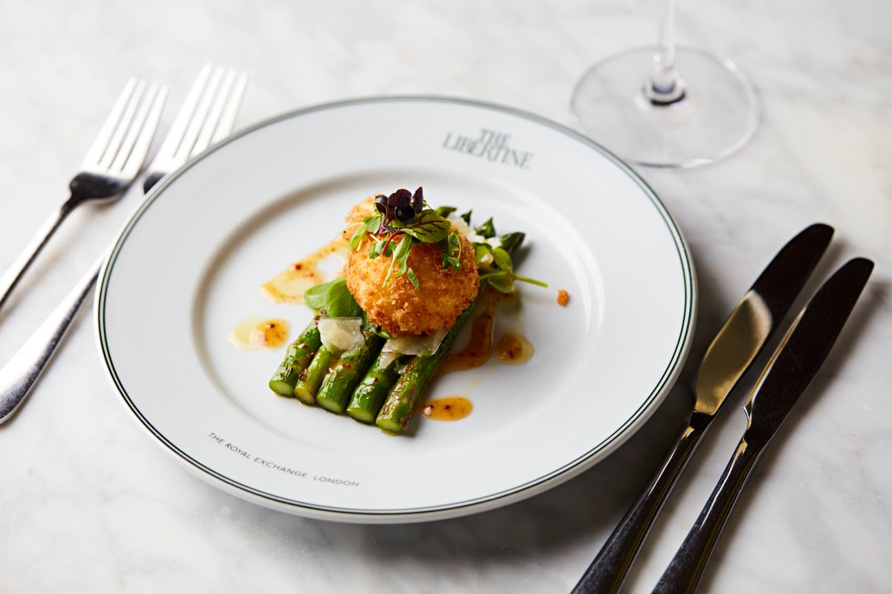 Scotch egg resting on asparagus on a plate in restaurant with The Libertine logo visible.